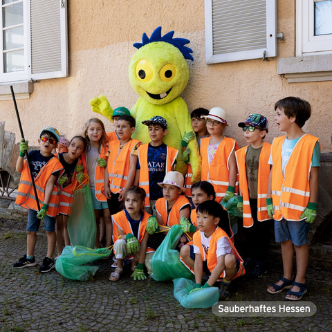 Bildtext:
Bildbeschreibung: Mehrere Schulkinder in Warnwesten und mit Müllsäcken zusammen mit dem Maskottchen Müllmo vor einer Wand.